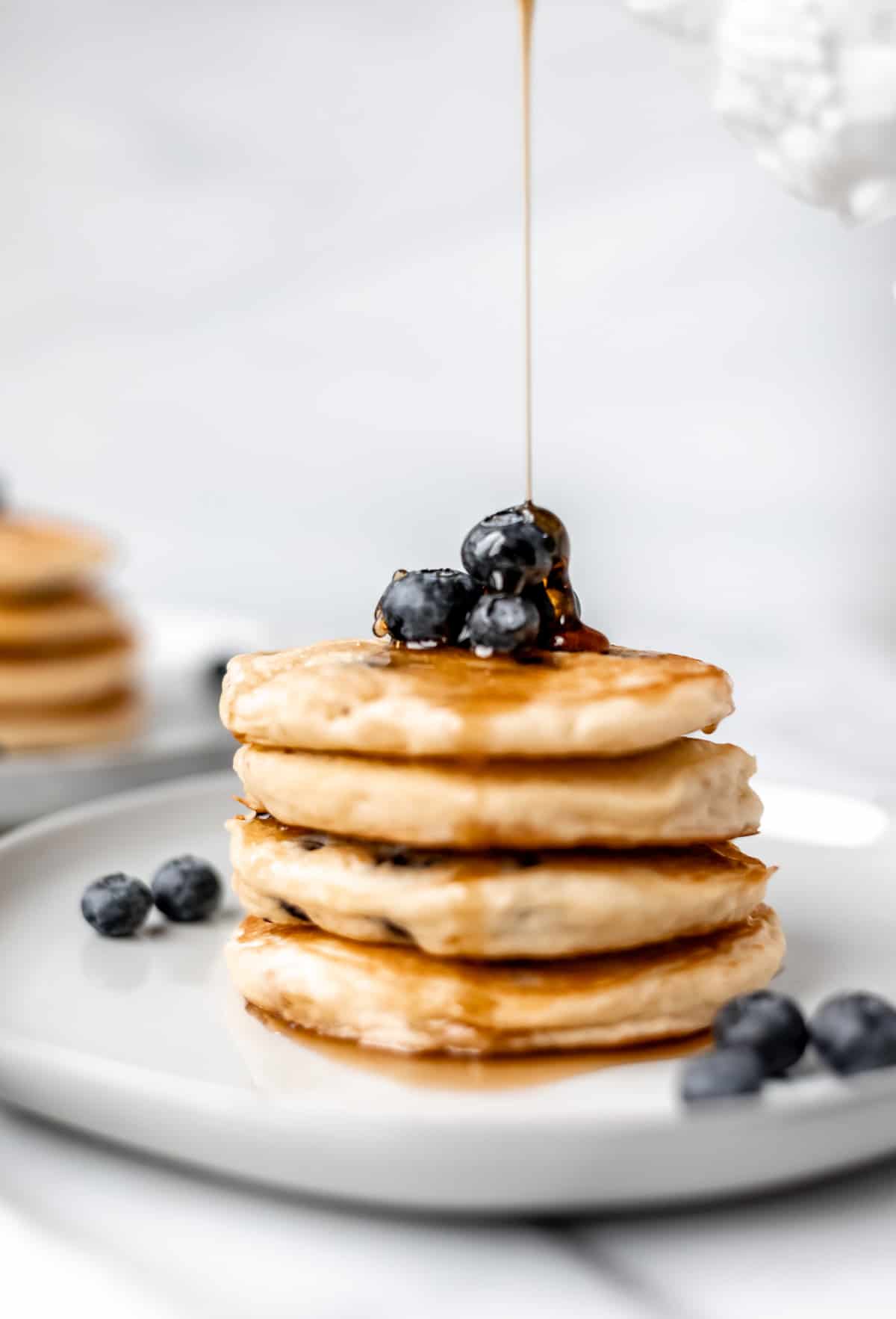 Maple syrup being poured over a stack of four fluffy blueberry pancakes on a white plate with extra blueberries and a second plate of pancakes in the background.