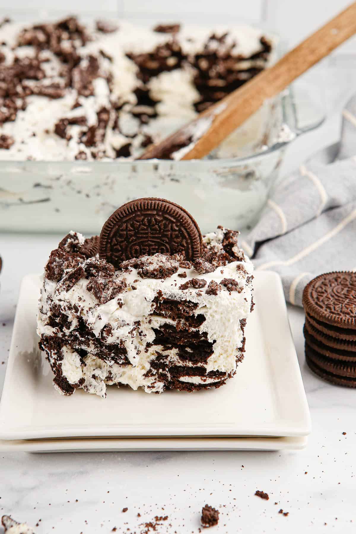 A serving of Oreo Icebox Cake on a white square plate with the baking dish of remaining cake in the background and a stack of Oreo cookies on one side.