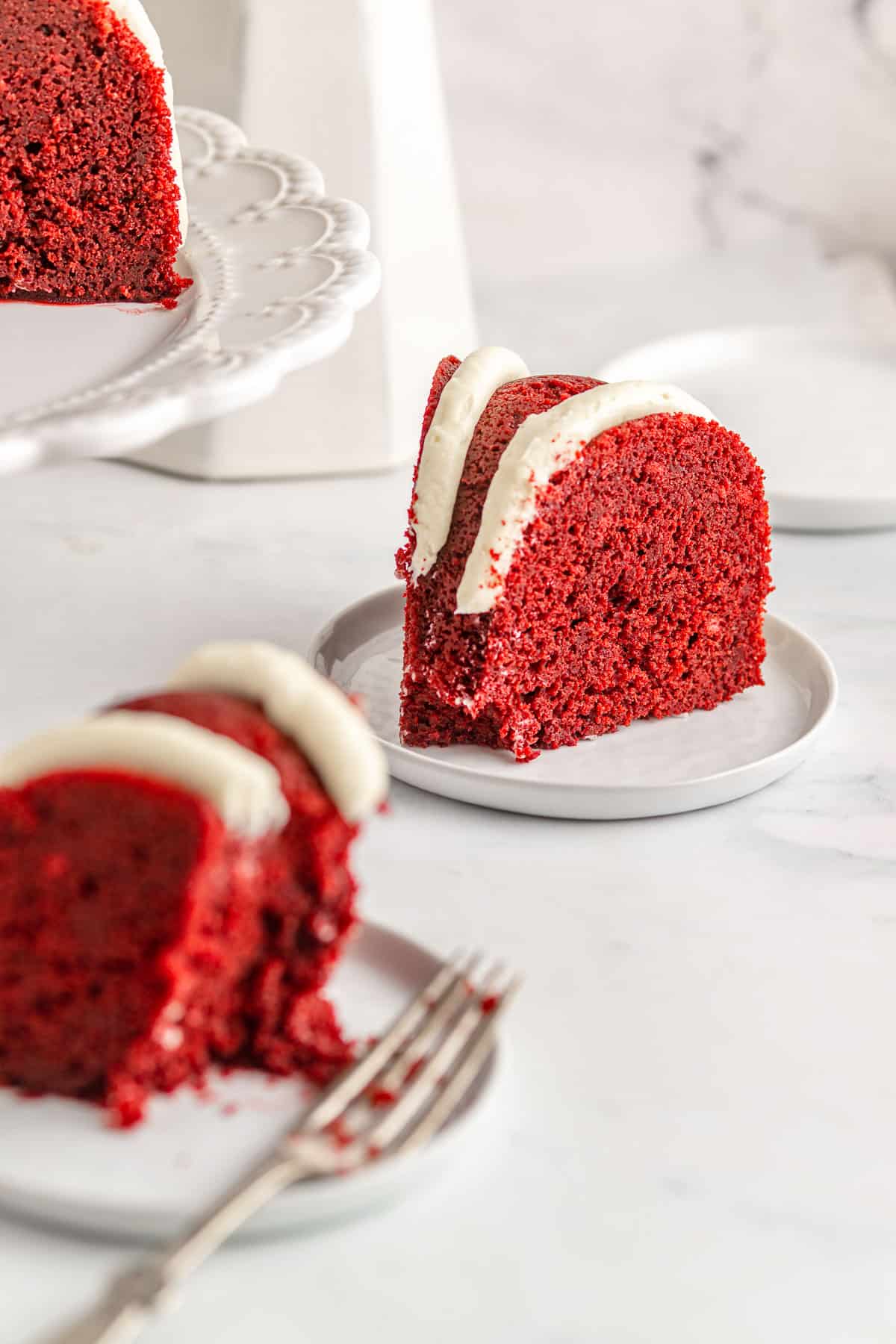 Two slices of red velvet pound cake with cream cheese frosting on white plates.