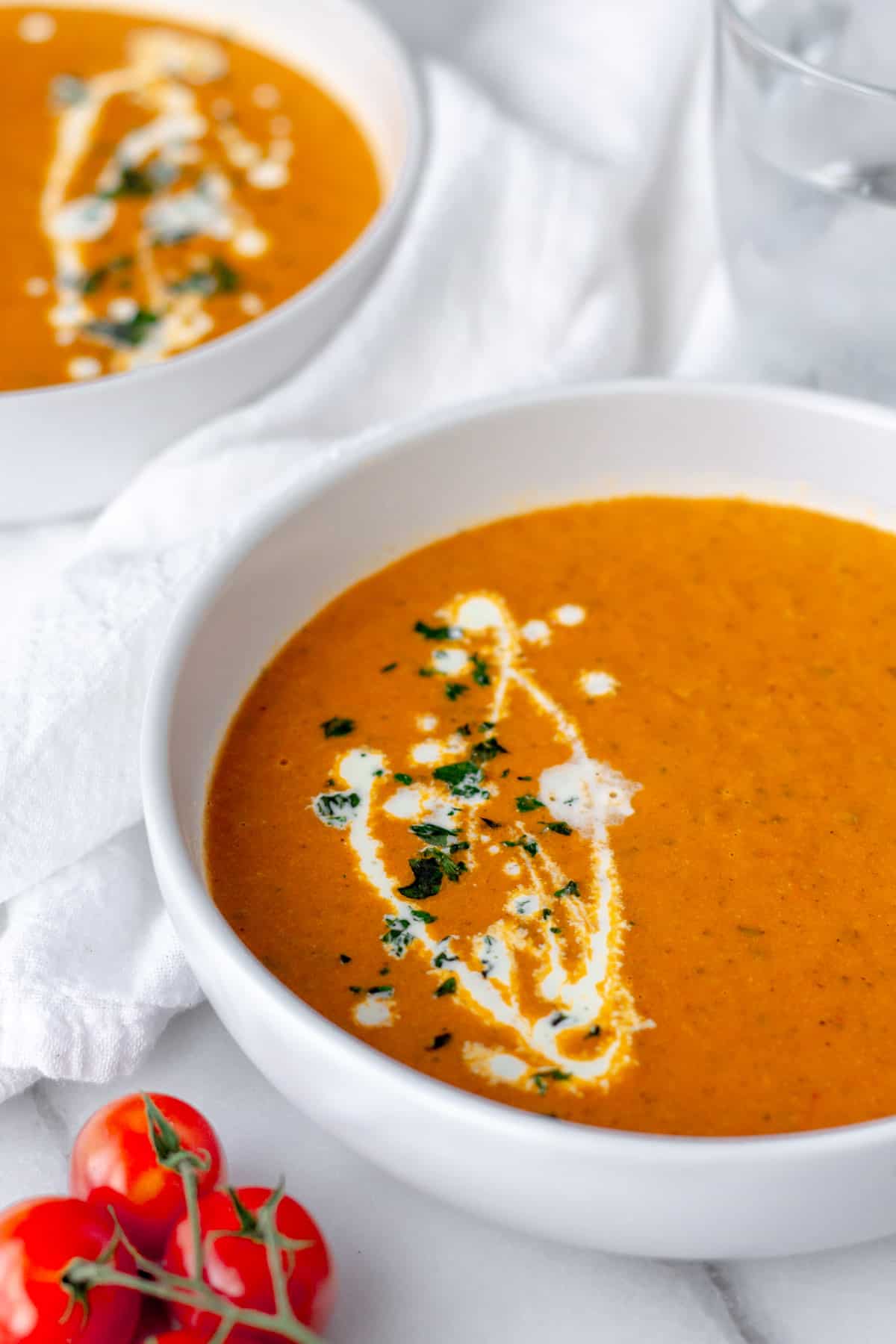 Two bowls of roasted tomato soup garnished with a drizzle of cream and basil with fresh cherry tomatoes in the foreground.