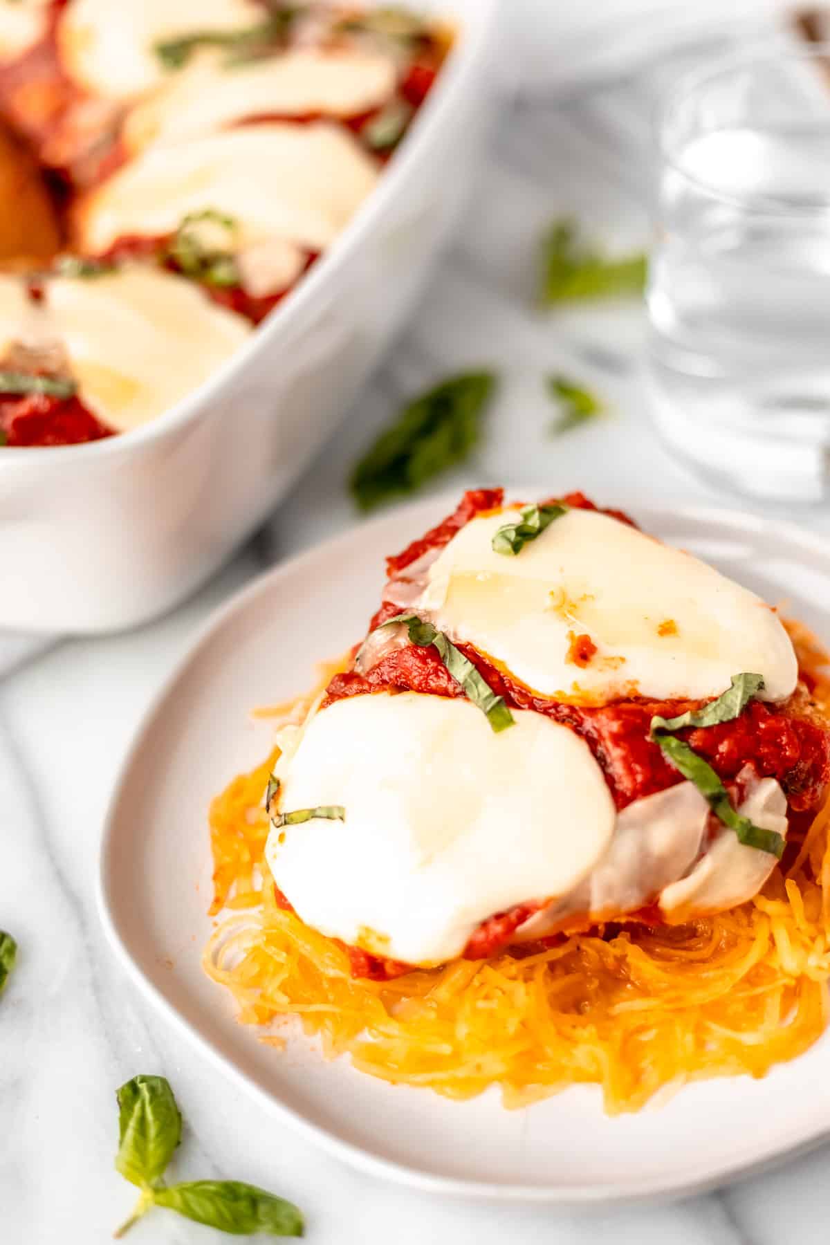 A serving of spaghetti squash chicken parmesan on a white dish with a white casserole dish and glass of water in the background.