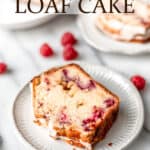 A slice of white chocolate raspberry loaf cake on a plate with a second serving and remaining loaf in the background with text overlay.