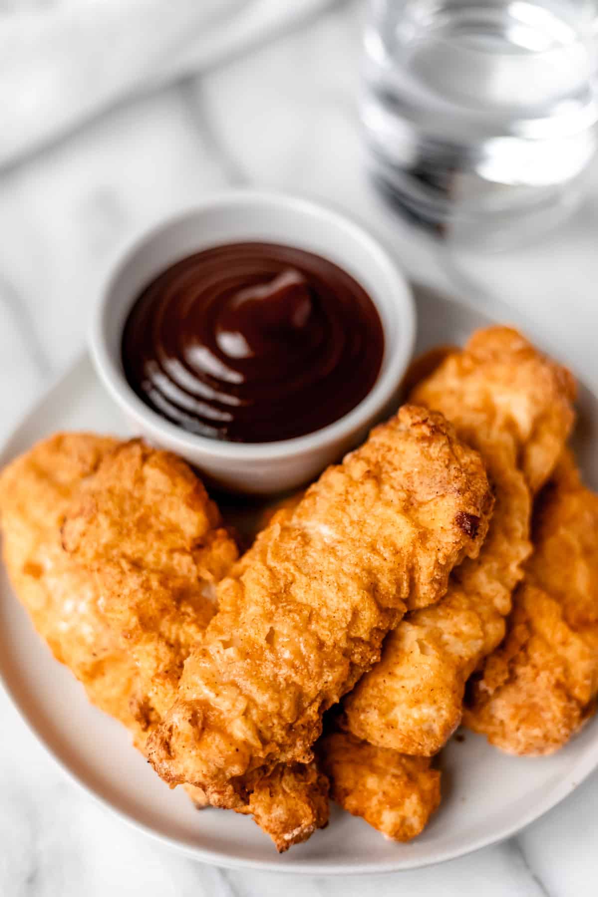 A stack of air fryer chicken tenders on a white plate with a white bowl of barbecue sauce and a glass of water in the background.