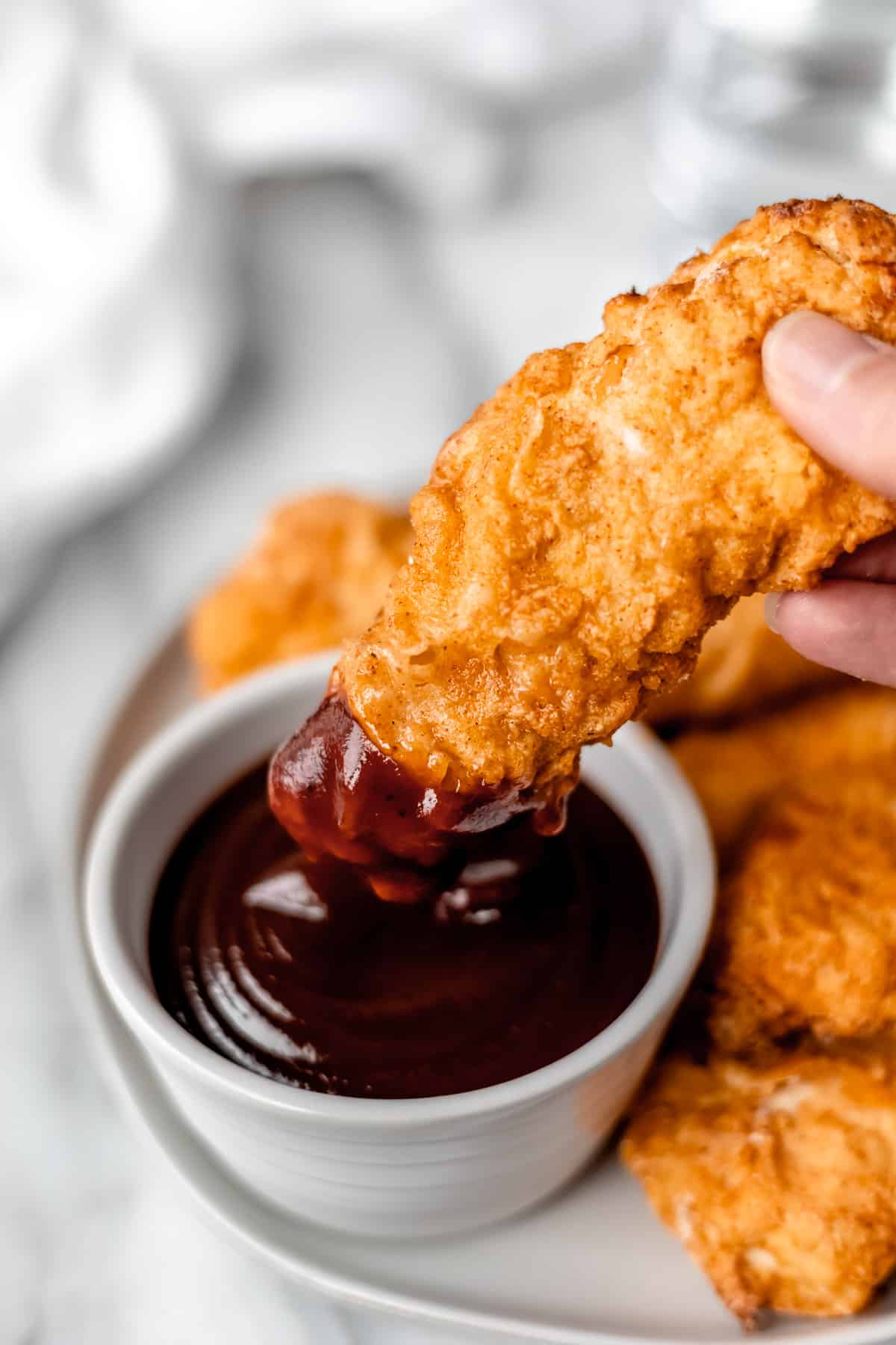 A chicken tender with barbecue sauce on one end being held up over the bowl of sauce.
