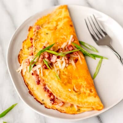 A ham and cheese omelet garnished with diced ham, cheese and green onions on a white plate with a fork.