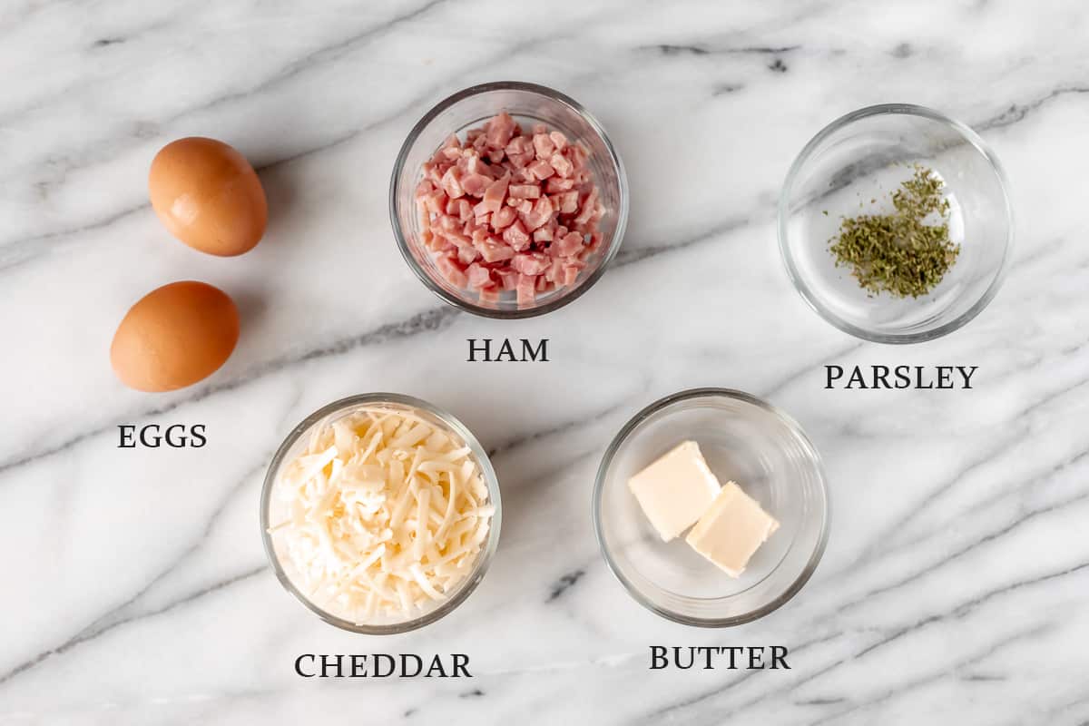 Ingredients needed to make a ham and cheese omelet on a marble background with text overlay.