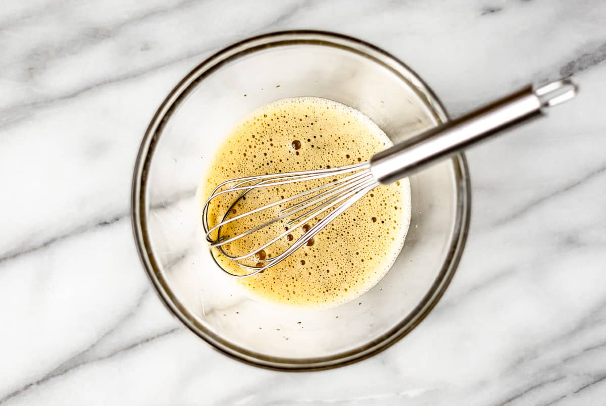 Whisked eggs with salt, pepper and parsley in a glass bowl with a silver whisk over a marble background.