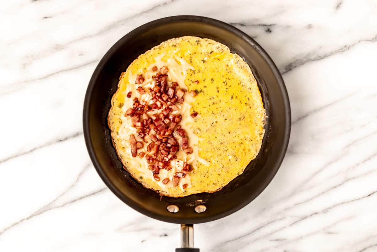 An omelet in the bottom of a pan with one half covered in cheese and diced ham over a marble background.