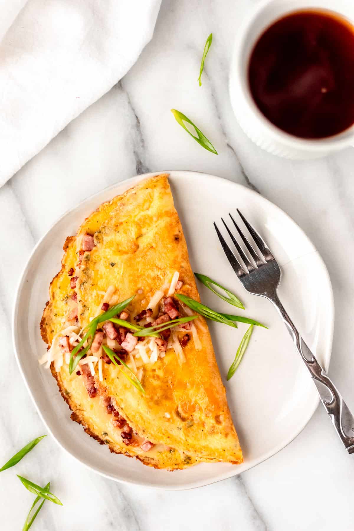 A ham and cheese omelet on a white plate garnished with diced ham, cheese and green onions with a fork on the black and cup of coffee and a white napkin around it.