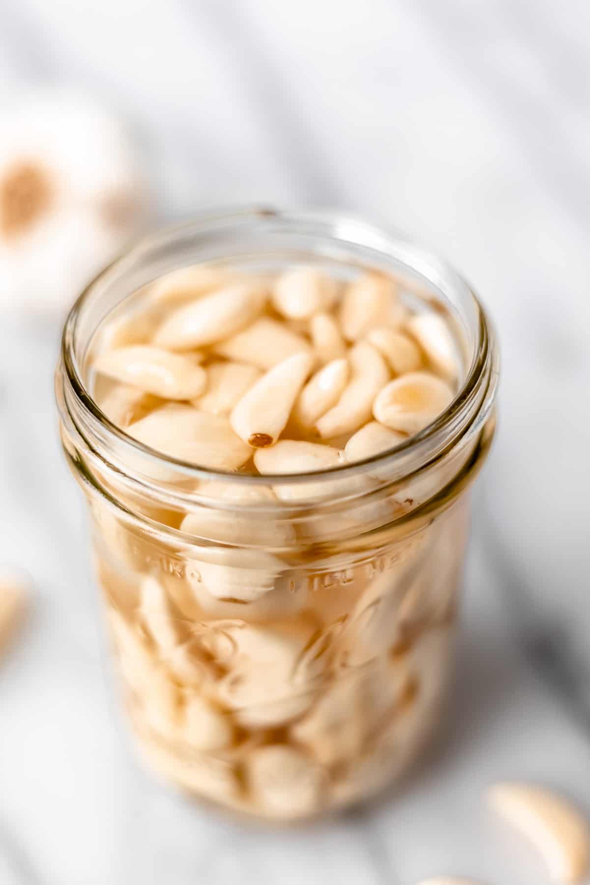 A jar full of pickled garlic with more cloves and a head of garlic around it.