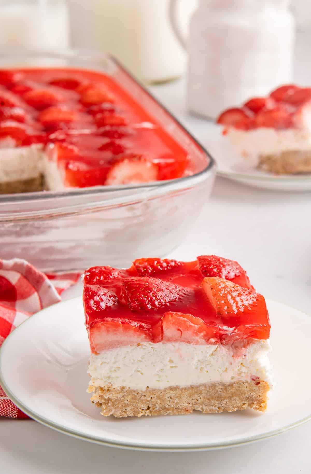 A slice of Strawberry Delight on a small plate with a second serving and a baking dish in the background.