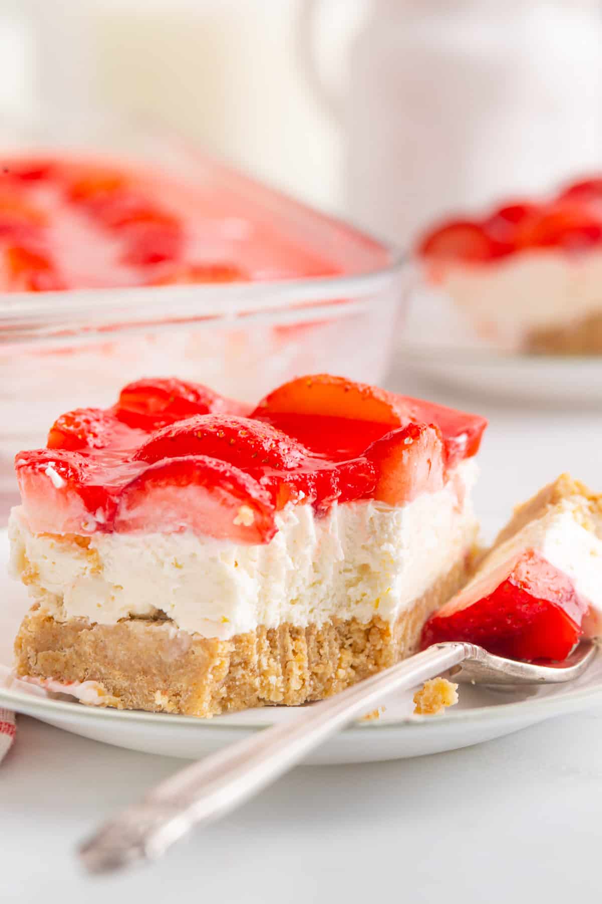 A slice of Strawberry Delight with a bite taken out on a fork on a small plate with a second serving and the baking dish in the background.
