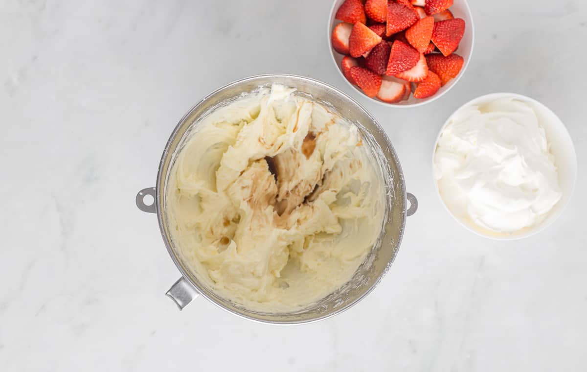 Cream cheese whipped in a mixing bowl with vanilla extract on top and a bowl of strawberries on the side.