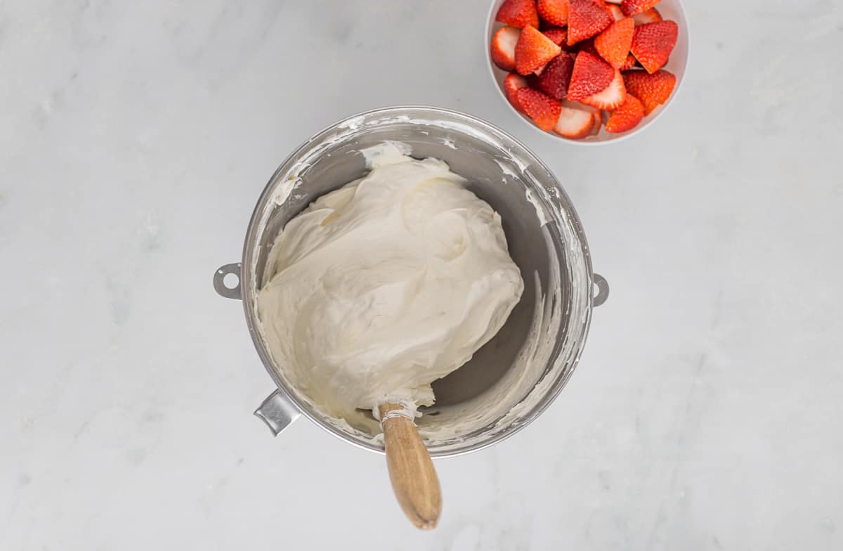 Cheesecake filling in a mixing bowl with a spatula and a bowl of strawberries off to the side.
