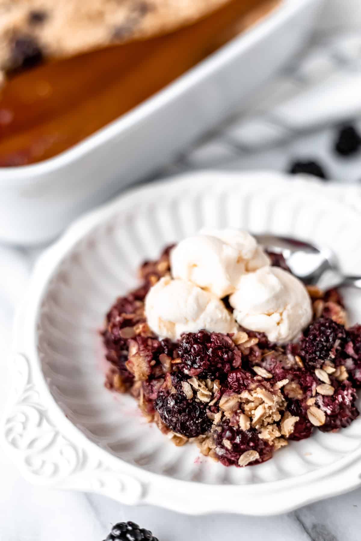 Blackberry crisp topped with vanilla ice cream in a white bowl with the baking dish partially showing in the background.