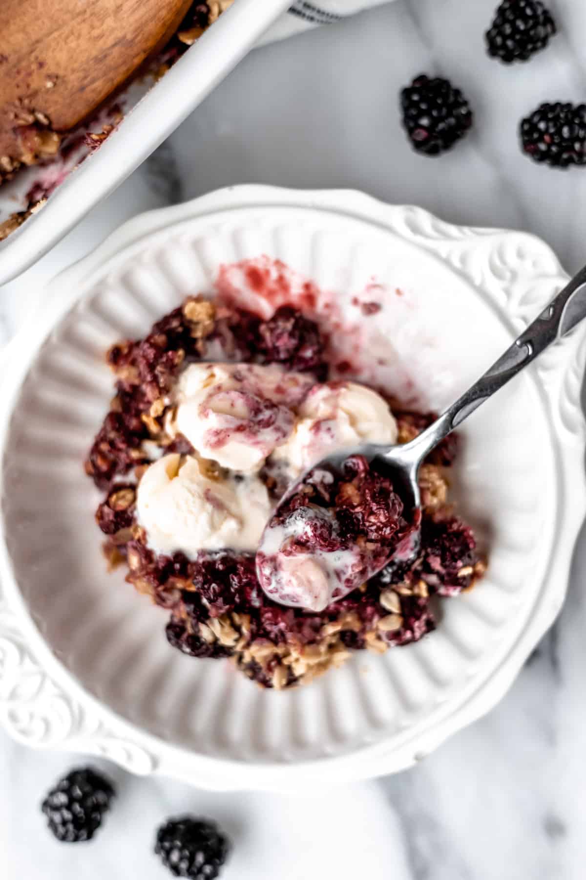 Overhead of blackberry crisp with ice cream with a scoop on a spoon in the bowl and blackberries around it.