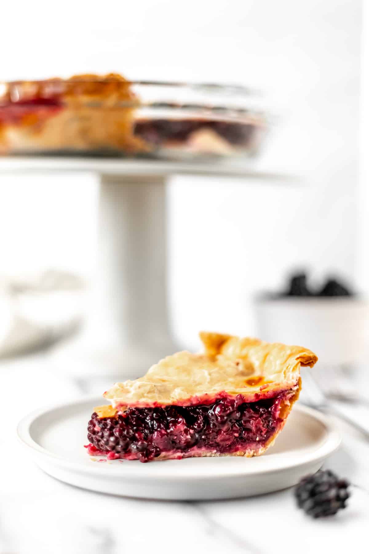 A slice of blackberry pie on a small plate with a cake stand and the remaining pie in the background with fresh blackberries around everything.