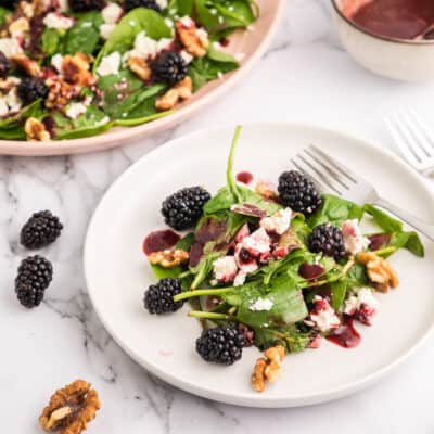 A blackberry salad on a white salad plate with the pink serving plate and bowl of blackberry vinaigrette in the background.