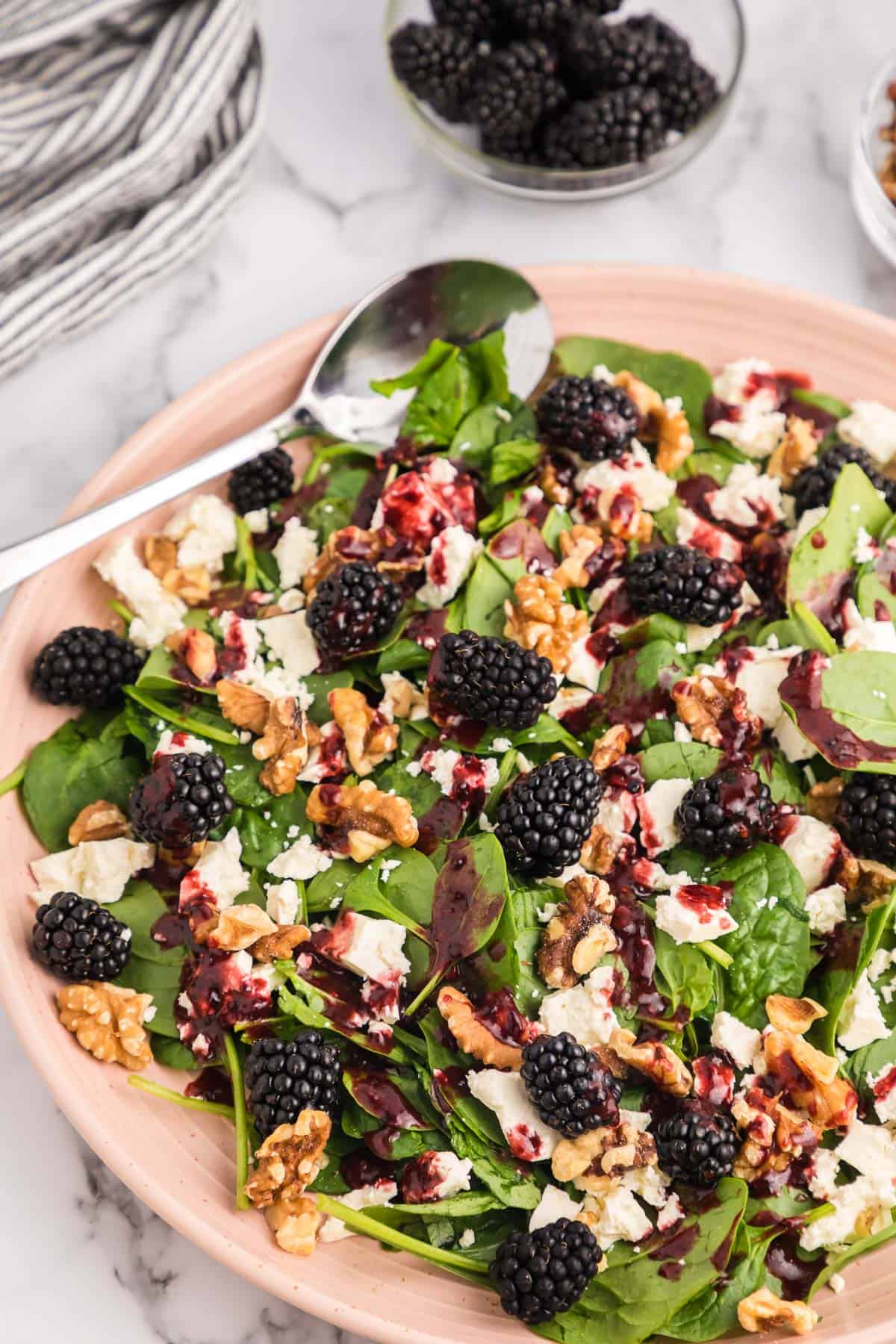 Spinach blackberry salad on a pink serving plate with a small bowl of fresh blackberries and a striped towel in the background.