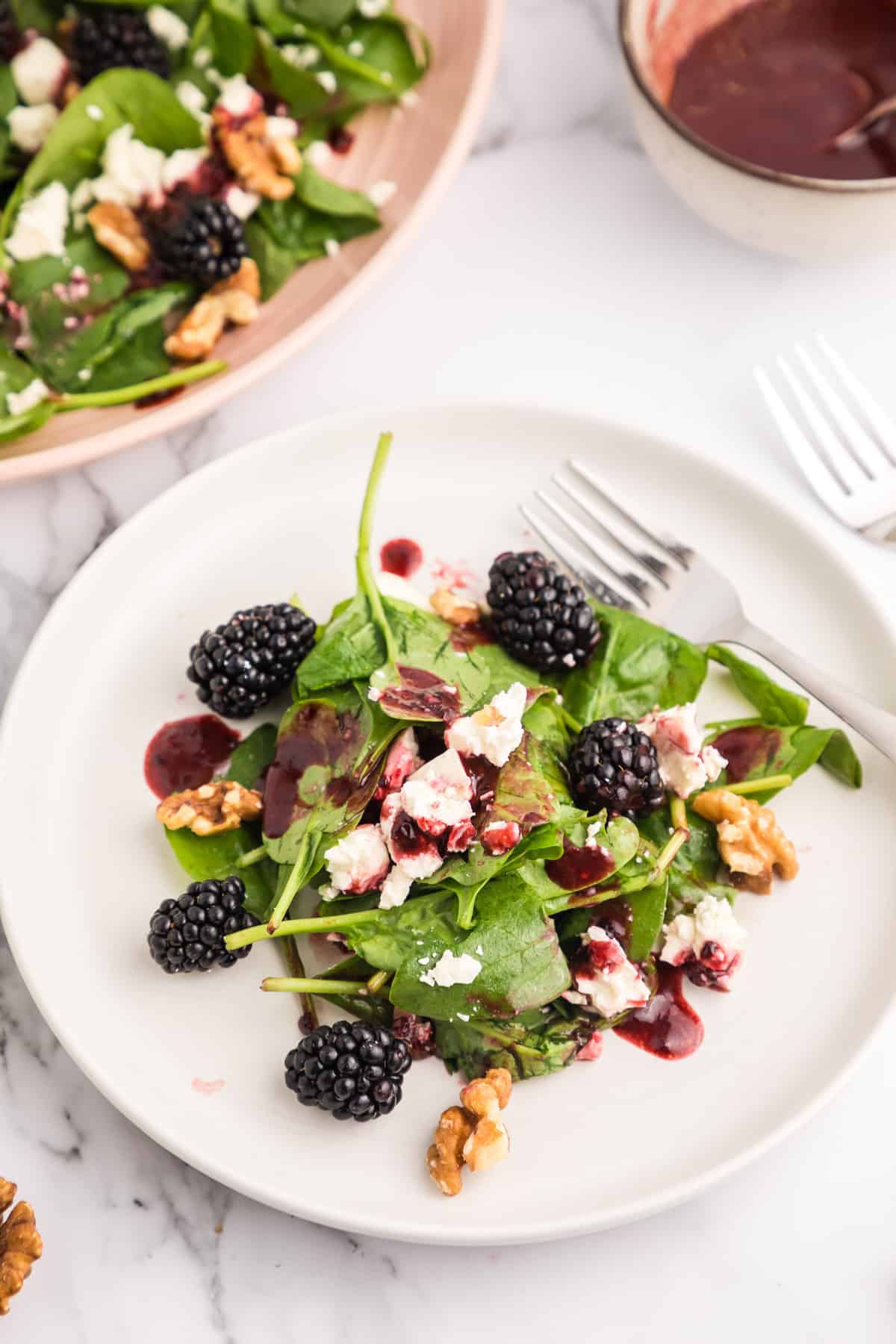 A blackberry salad on a white salad plate with the pink serving plate and bowl of blackberry vinaigrette in the background.