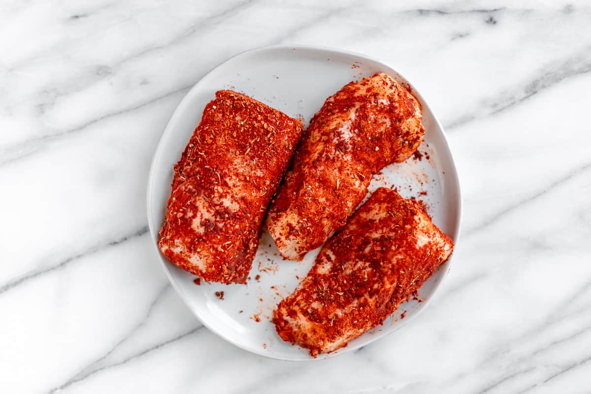 Three cod fillets coated in blackening seasoning on a white plate over a marble background.