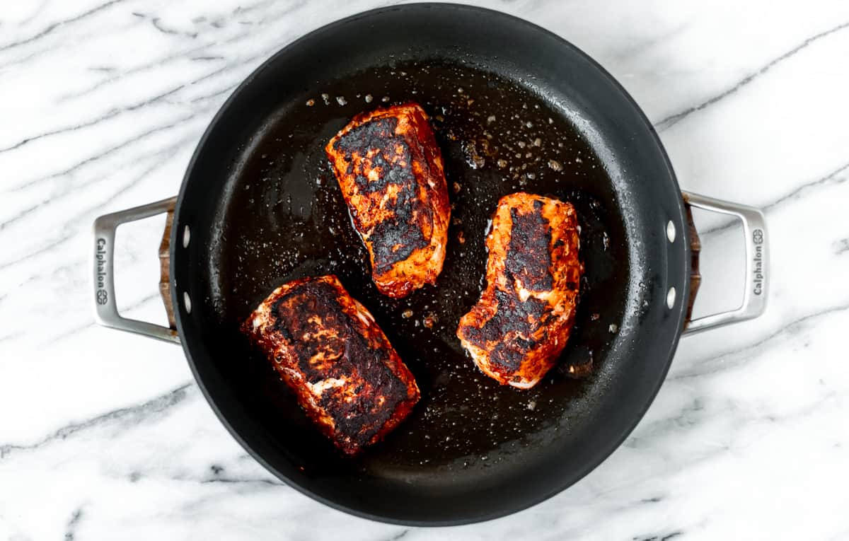 Three cooked blackened cod fillets in a black skillet over a marble background.