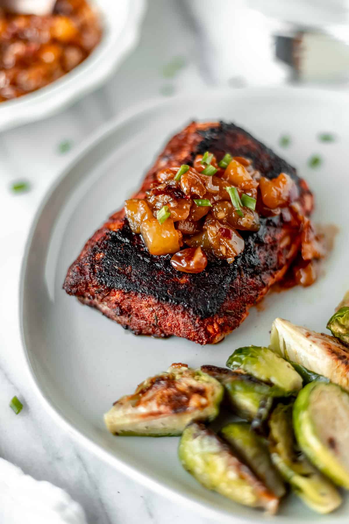 A blackened cod fish fillet topped with peach chutney on a white plate with brussels sprouts.