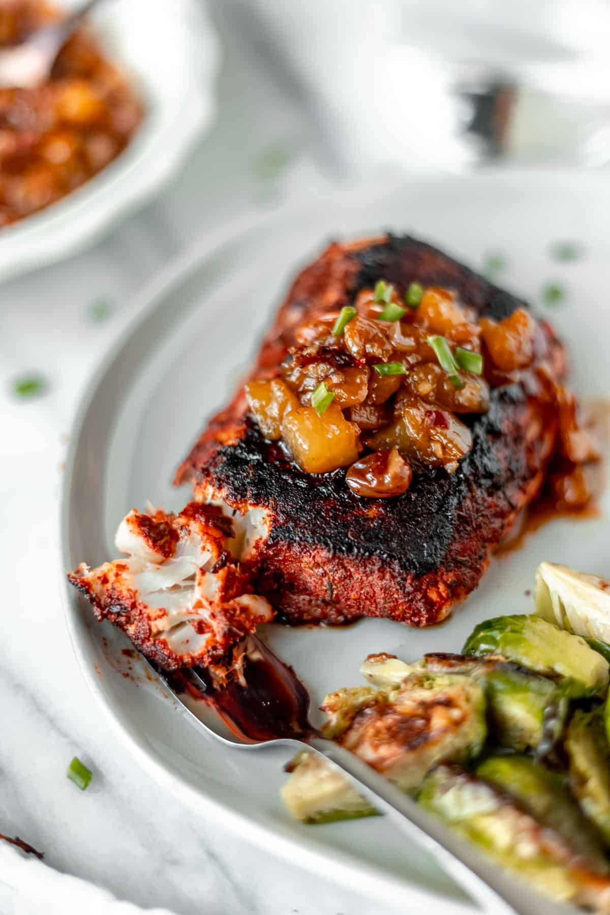 A blackened cod fillet with a piece cut off on a fork on a plate with brussels sprouts.