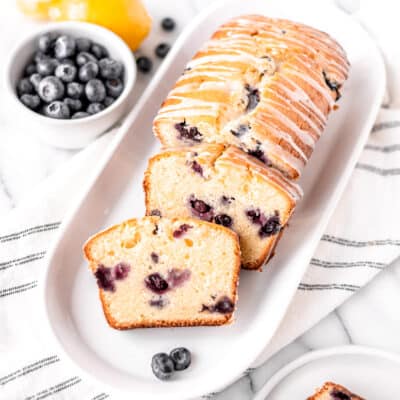 A lemon blueberry pound cake with several slices taken out on a serving plate with a striped tower and bowl of blueberries and lemon in the background.