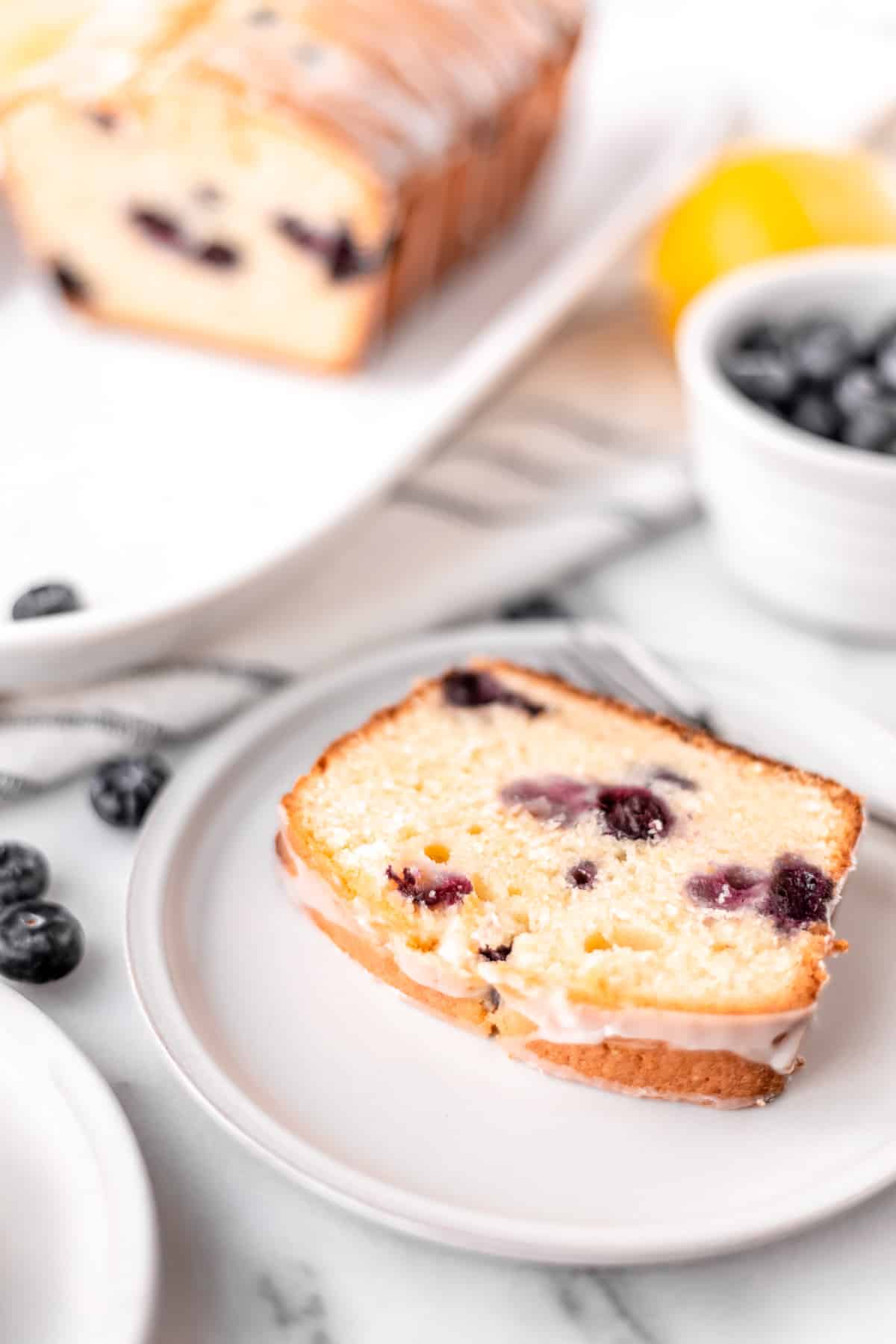 A slice of lemon blueberry pound cake on a small white plate with the remaining cake, a bowl of blueberries and a lemon in the background.