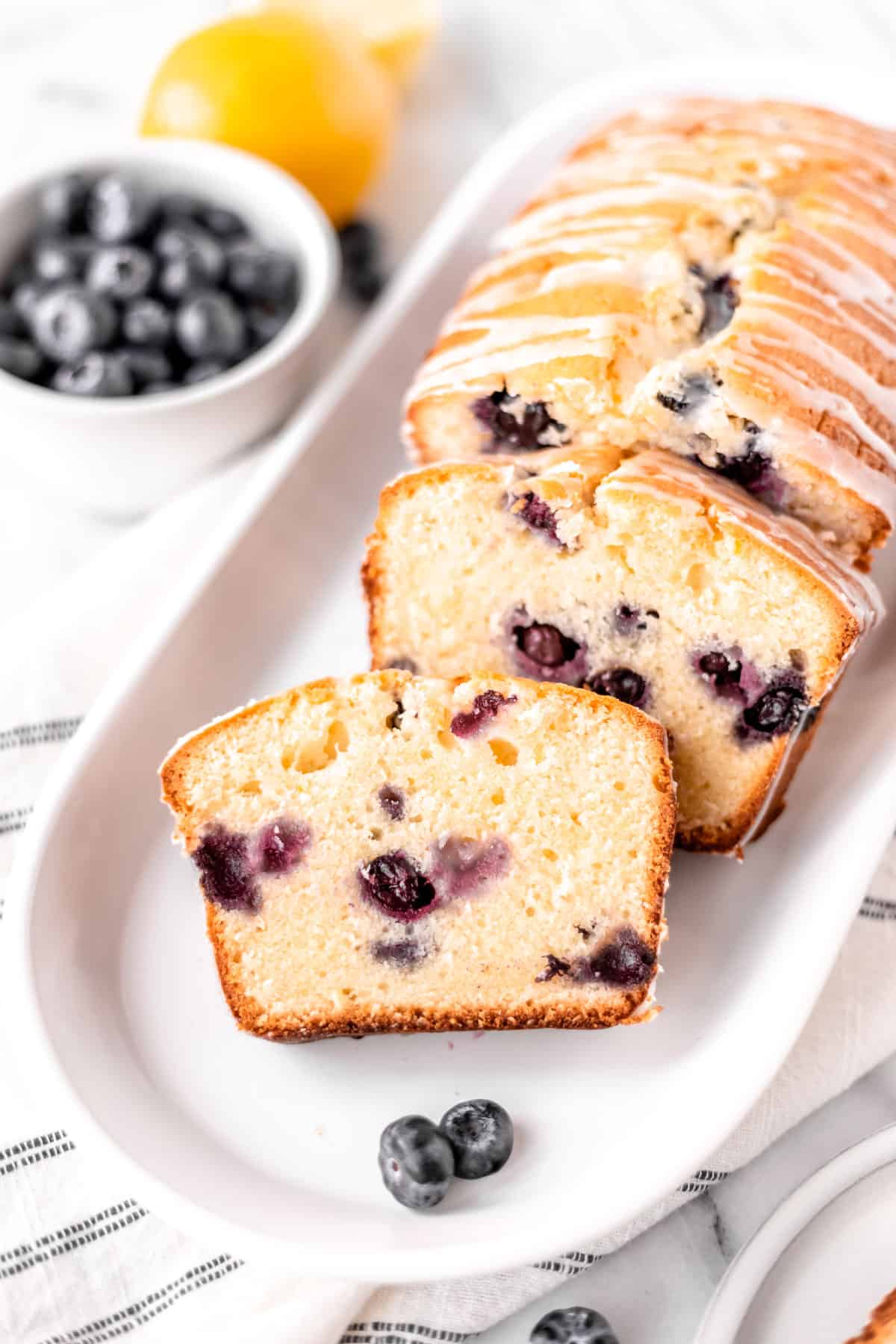 A lemon blueberry pound cake with several slices taken out on a serving plate with a striped tower and bowl of blueberries and lemon in the background.