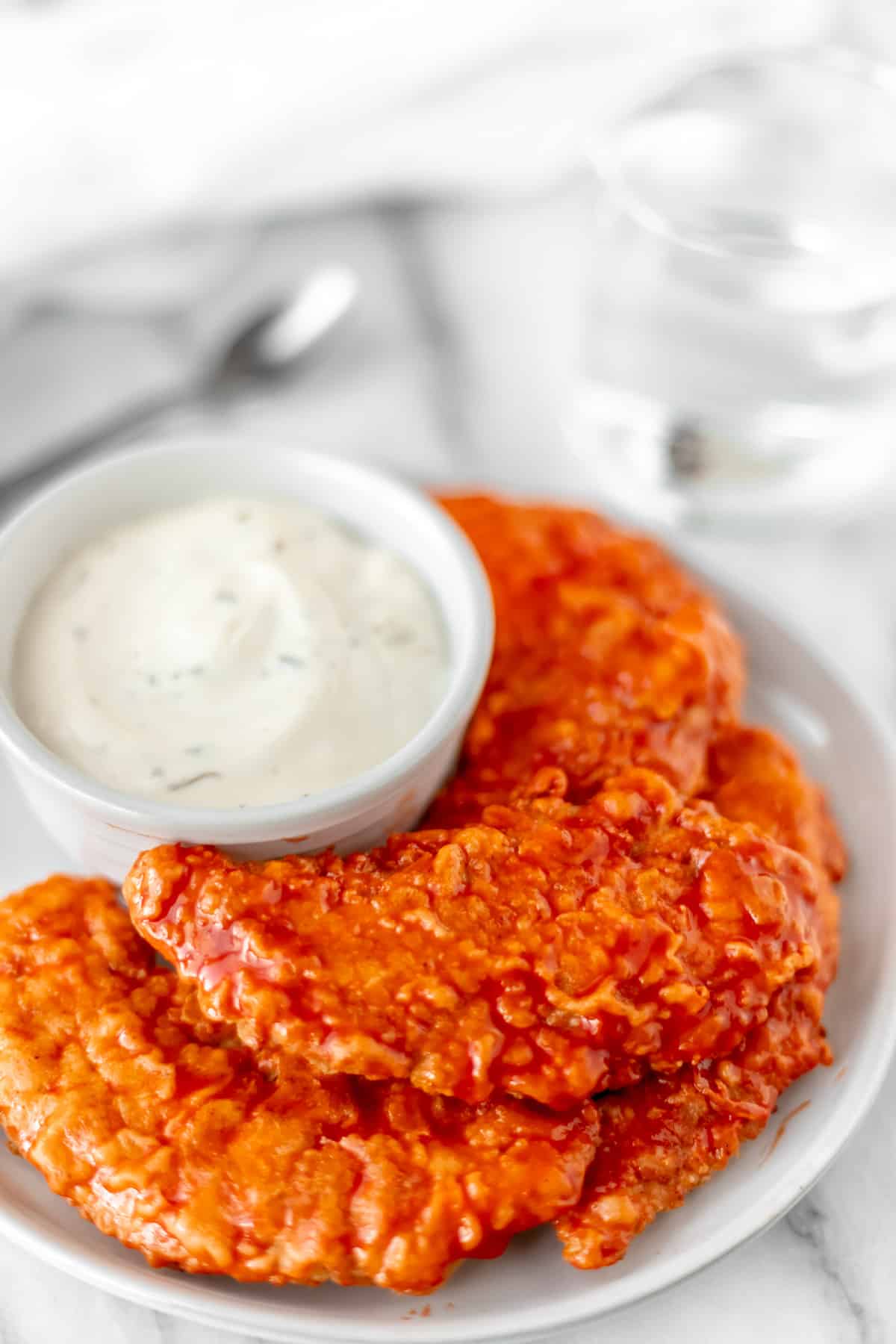 Buffalo chicken tenders stacked on a white plate with a white bowl of ranch dressing.