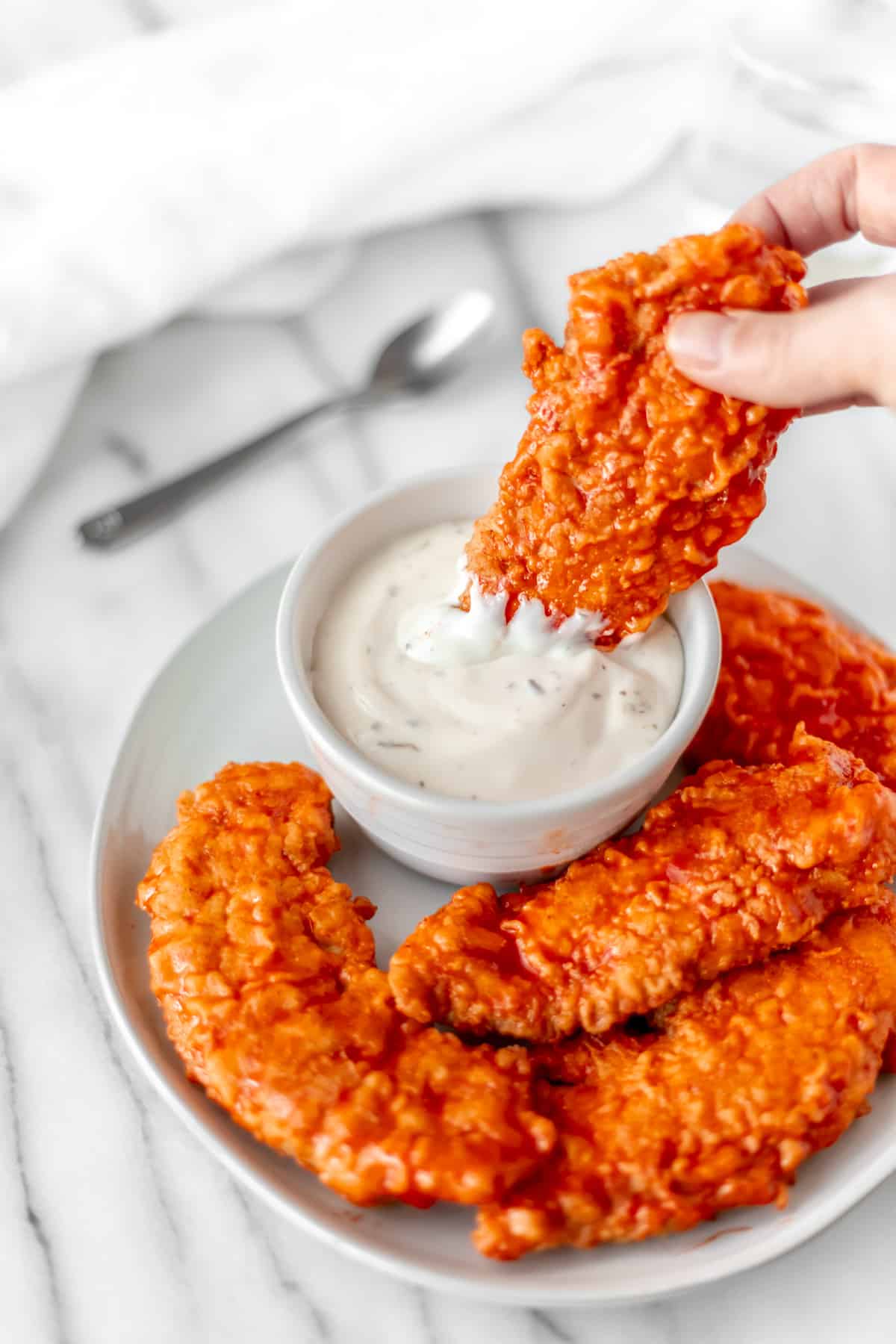 A buffalo chicken tender being dipped into ranch dressing that's in a white bowl on a white plate with more buffalo chicken tenders.