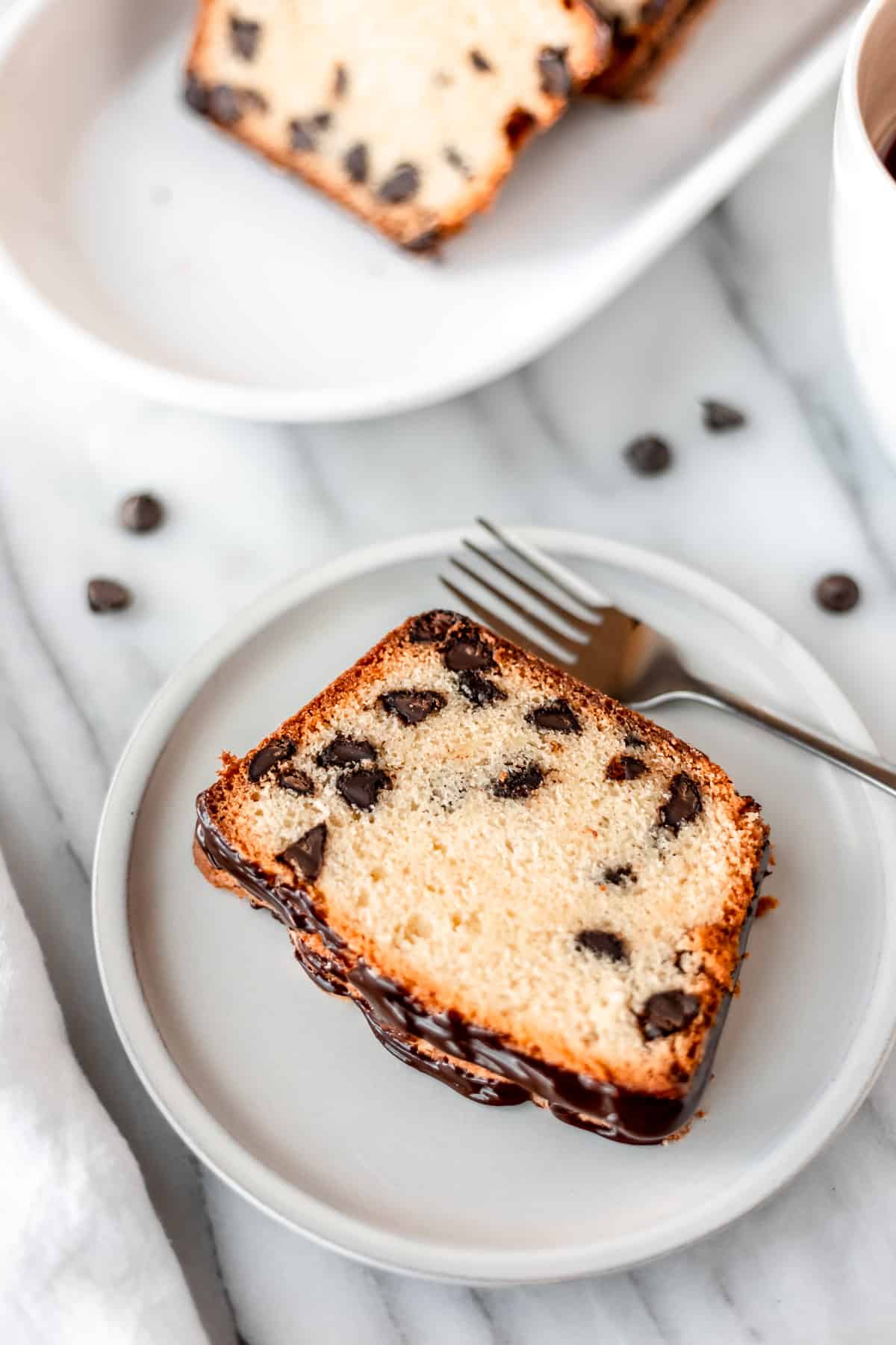 A slice of chocolate chip pound cake on a small white plate with a fork with chocolate chips and a serving tray of more pound cake partially showing around it.
