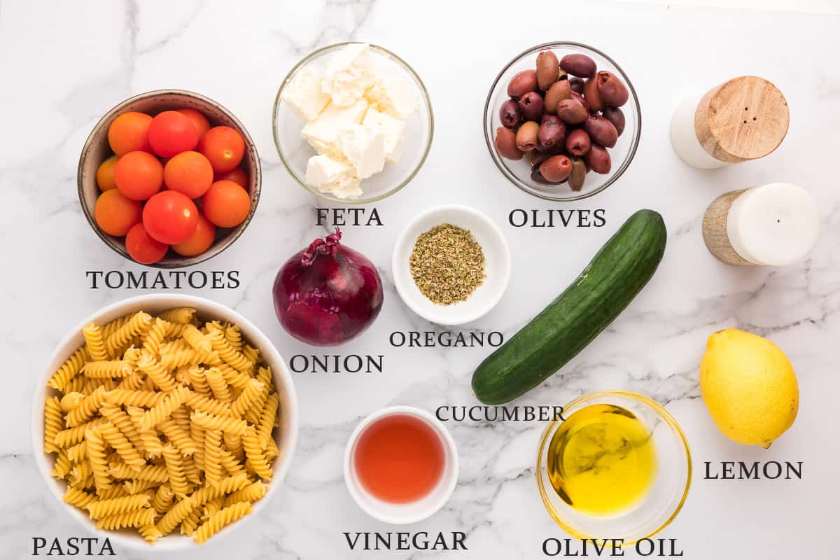 Ingredients needed to make Greek pasta salad on a marble background with text overlay.