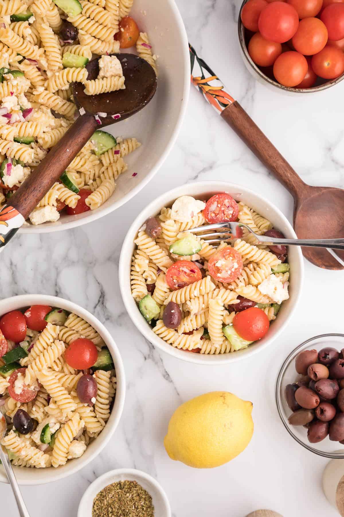 Overhead of two bowls of Greek pasta salad with a large bowl and small bowls of extra ingredients around them.