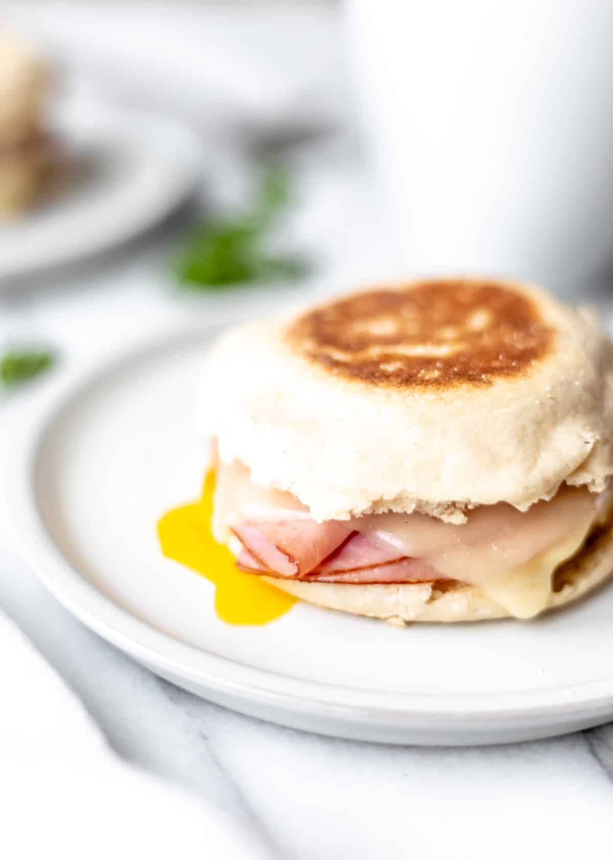 A ham, egg and cheese breakfast sandwich on a plate with yolk dripping out and parsley, a mug and part of a second plate in the background.