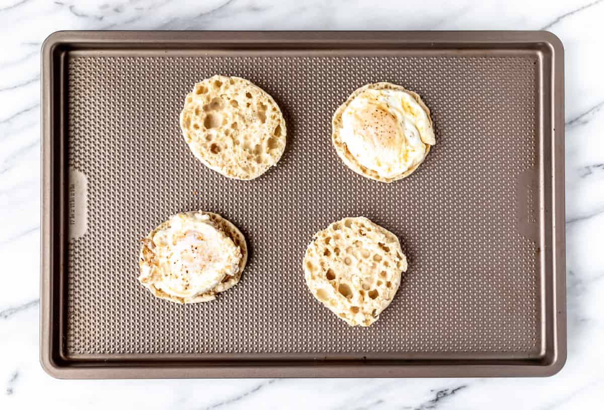 Four English muffin halves with eggs on top of two of them on a baking sheet.