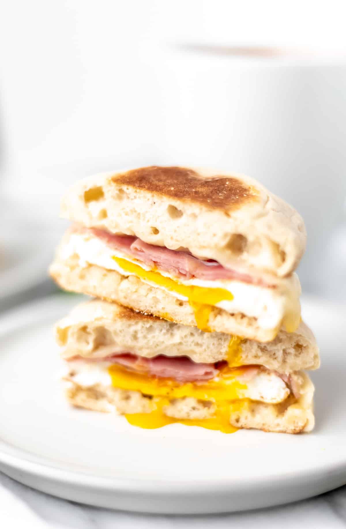 Two halves of an egg, ham, and cheese sandwich on an English muffin stacked on top of each other with yolk dripping down onto the plate.
