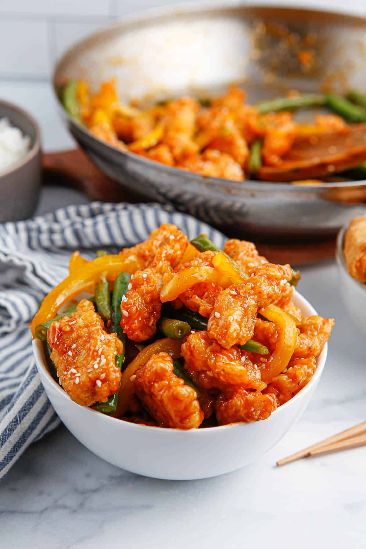 A bowl of copycat Panda Express Honey Sesame Chicken with a towel and a skillet with more chicken in it in the background.