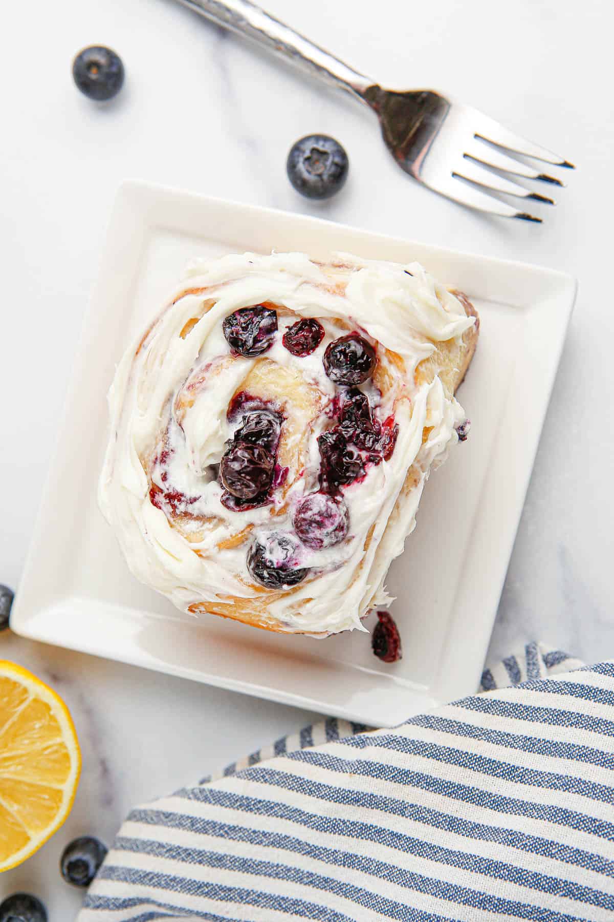 Overhead of a lemon blueberry sweet roll on a white plate with a lemon half, fork and striped towel around it.