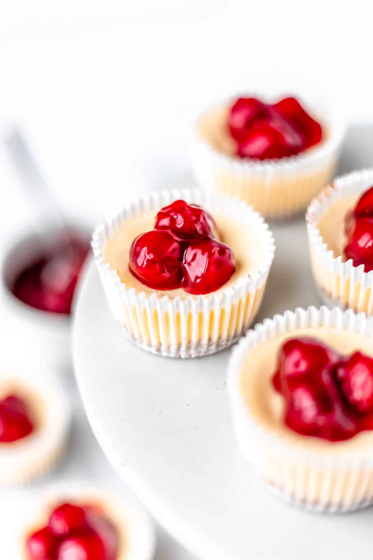 Mini cheesecake cupcakes topped with cherry pie filling on a cake stand with more on the table below.