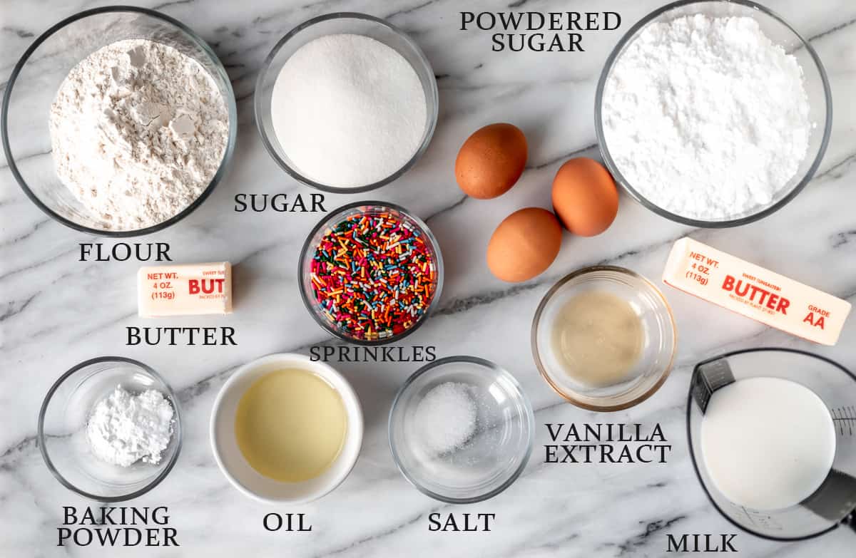 Ingredients needed to make mini sprinkle cupcakes on a marble background with text overlay.