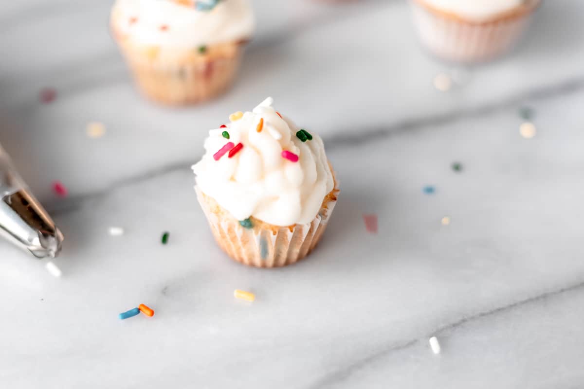 Rainbow sprinkles being dropped on top of a mini sprinkle cupcake.