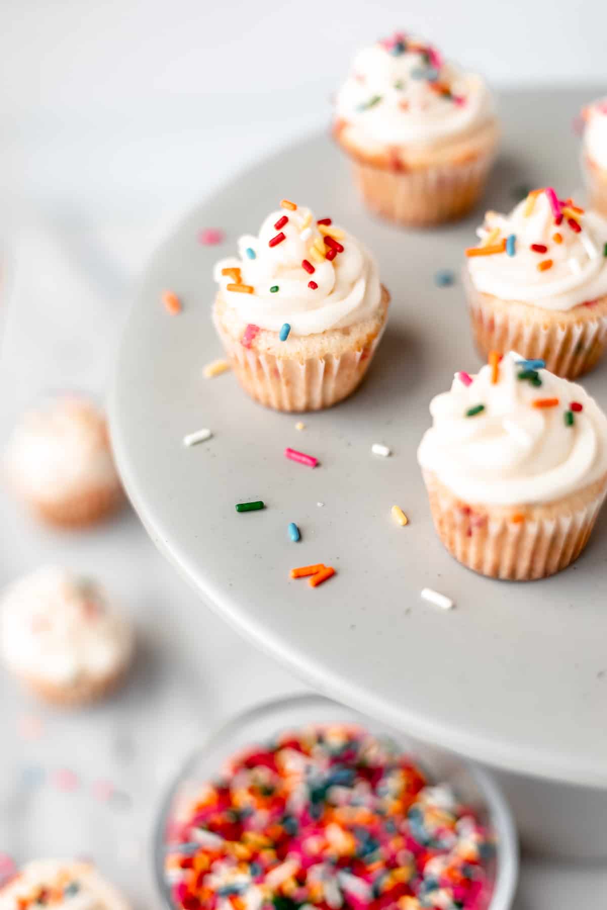 Mini sprinkle cupcakes on a gray cake stand with extra sprinkles around them and more cupcakes and a bowl of sprinkles below.