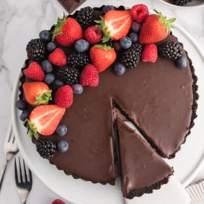 Overhead of a chocolate tart topped with fresh berries with a slice being lifted out on a pie server with forks, chocolate and berries around it.