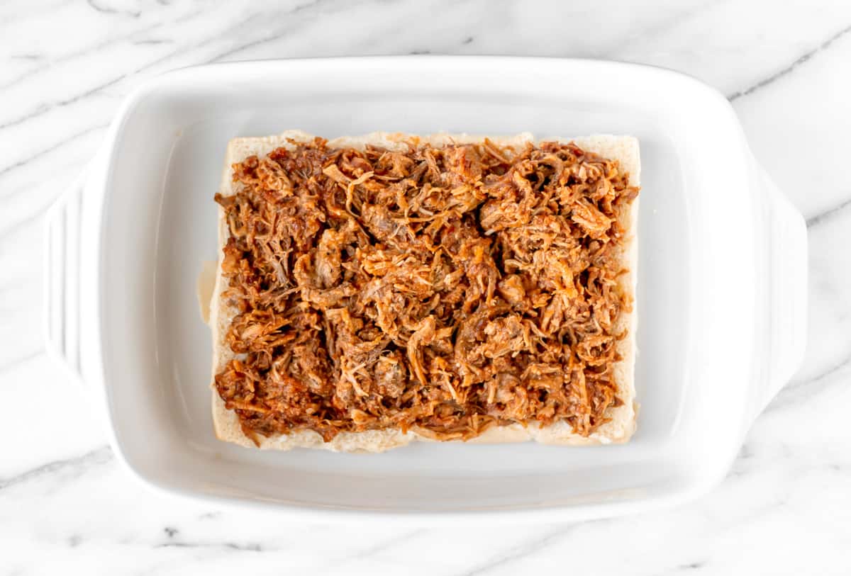 The bottom half of a pack of King's Hawaiian Rolls topped with pulled pork in a white casserole dish.