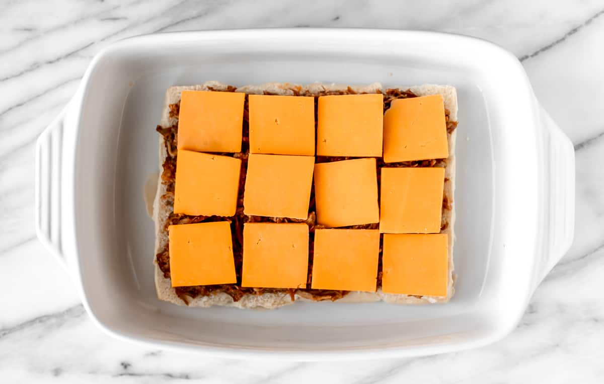 Slices of cheddar cheese on top of pulled pork on rolls in a casserole dish.