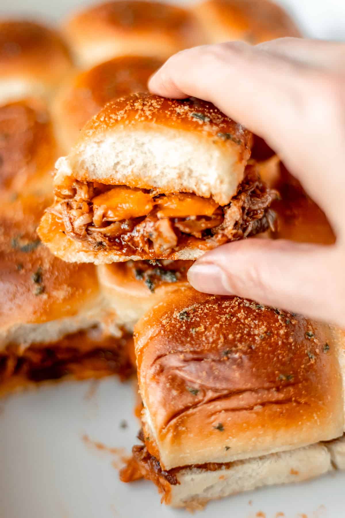 A hand holding a pulled pork slider on a stack of other pulled pork sliders.