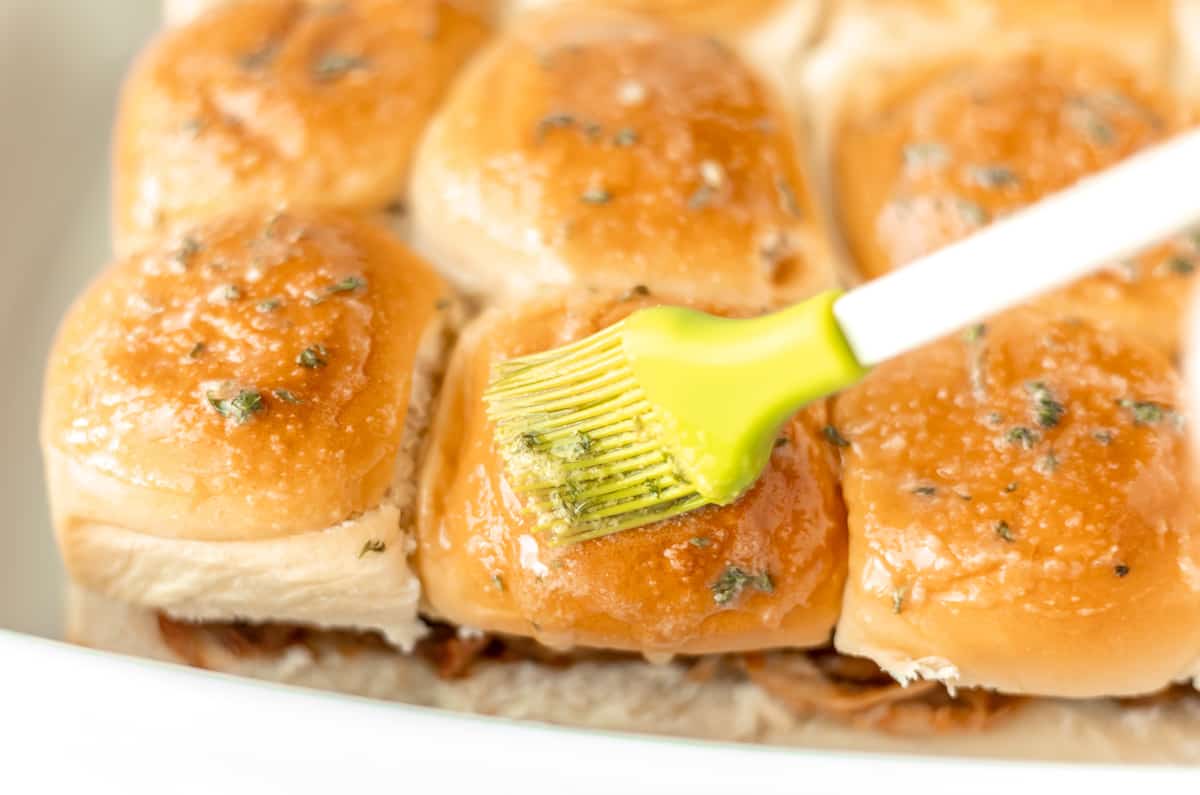 Garlic butter being brushed onto the tops of pulled pork sliders.